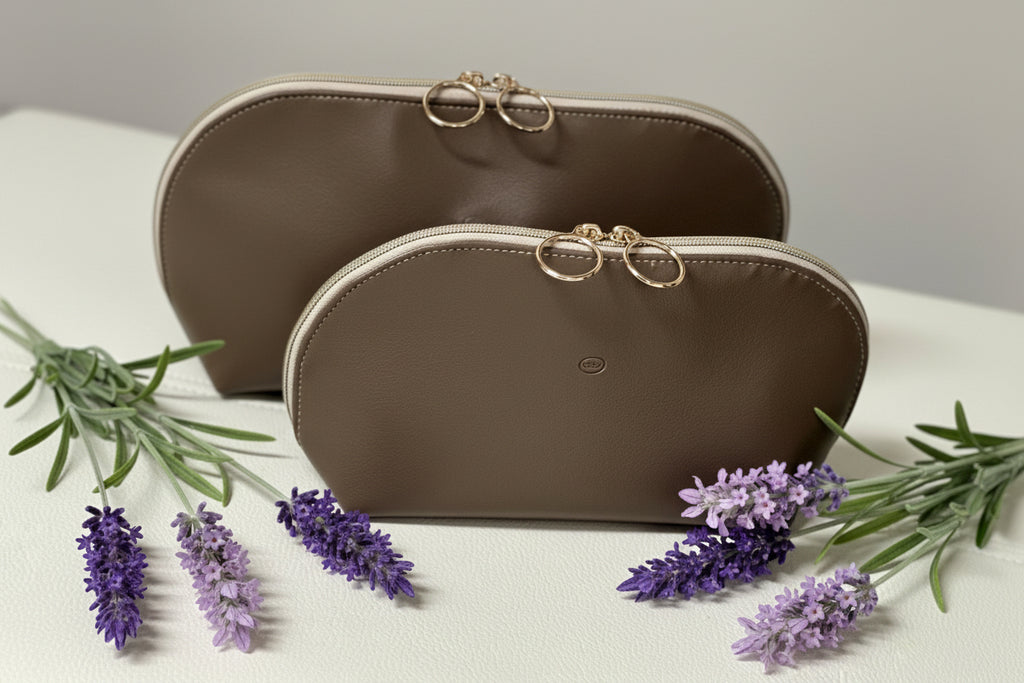 Two brown cosmetic bags with gold zippers on a white surface with lavender flowers.
