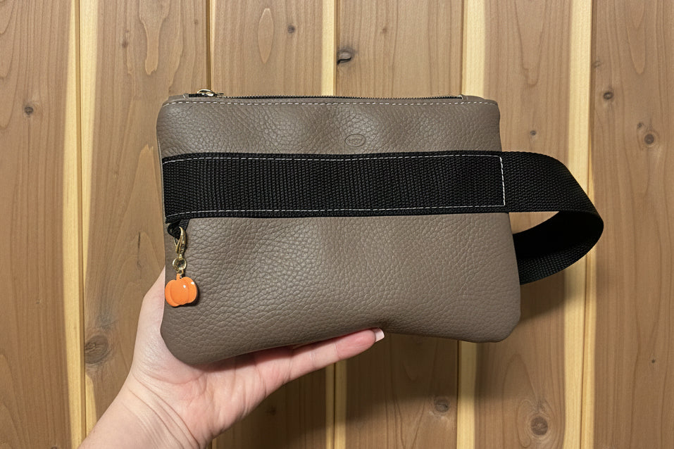 Brown leather wrist bag with black strap held against a wooden background