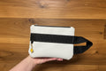 White wrist bag with a black strap held by a hand on a wooden surface