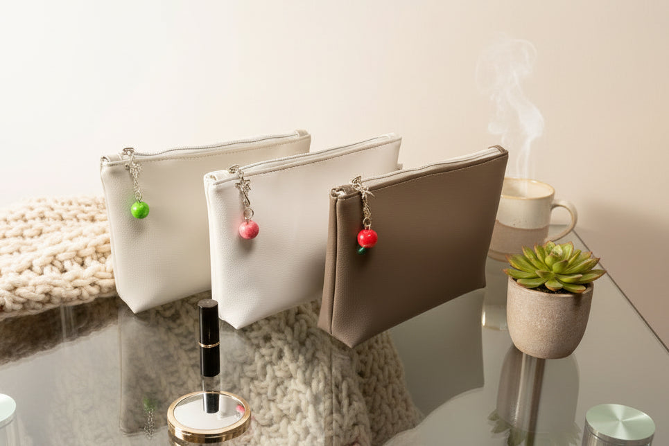 Three faux leather cosmetic bags with colorful individual charms on a reflective surface with a plant and candle in the background.