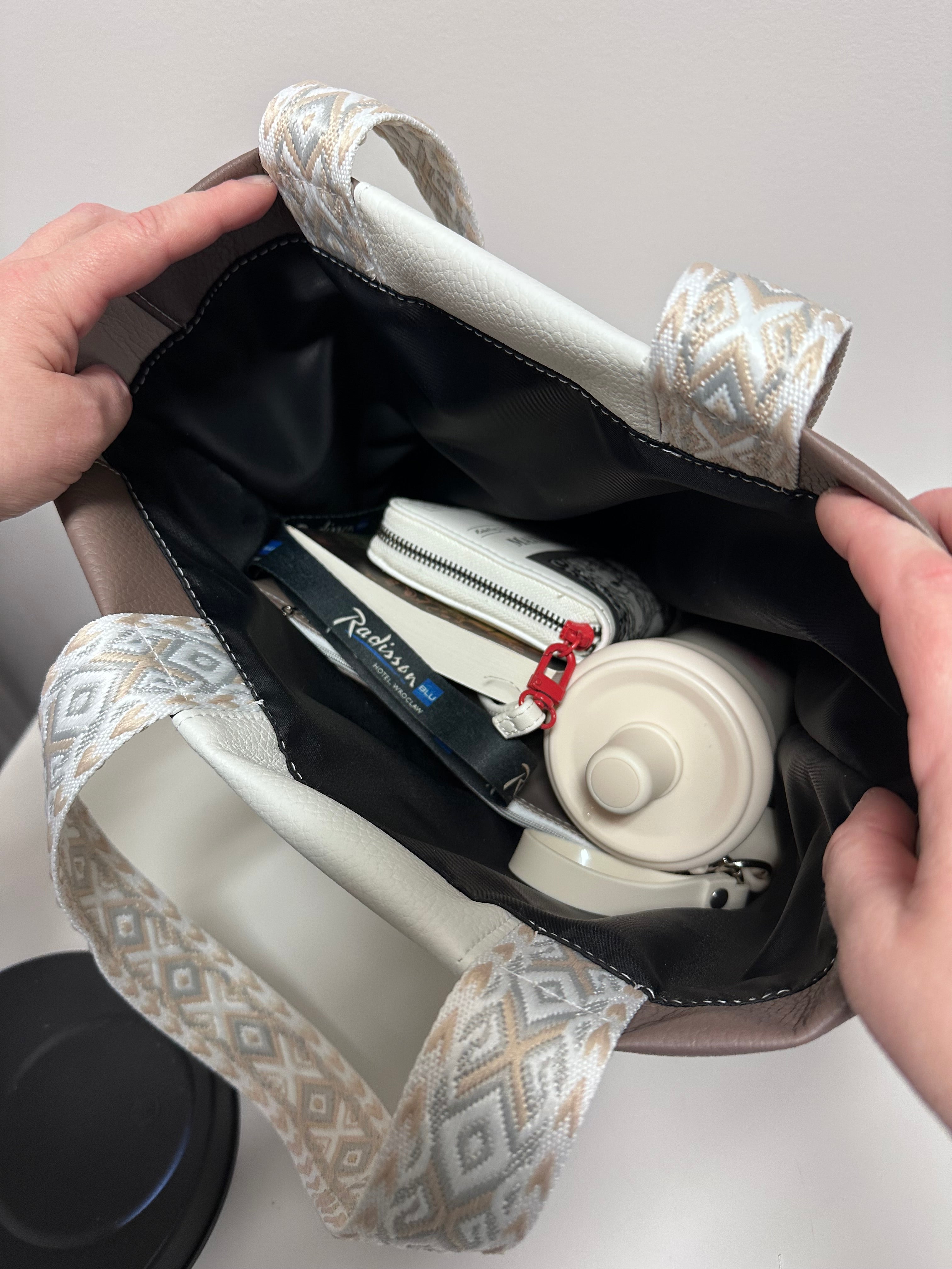 Handbag with a white handbag and small white container inside, held by hands against a neutral background.