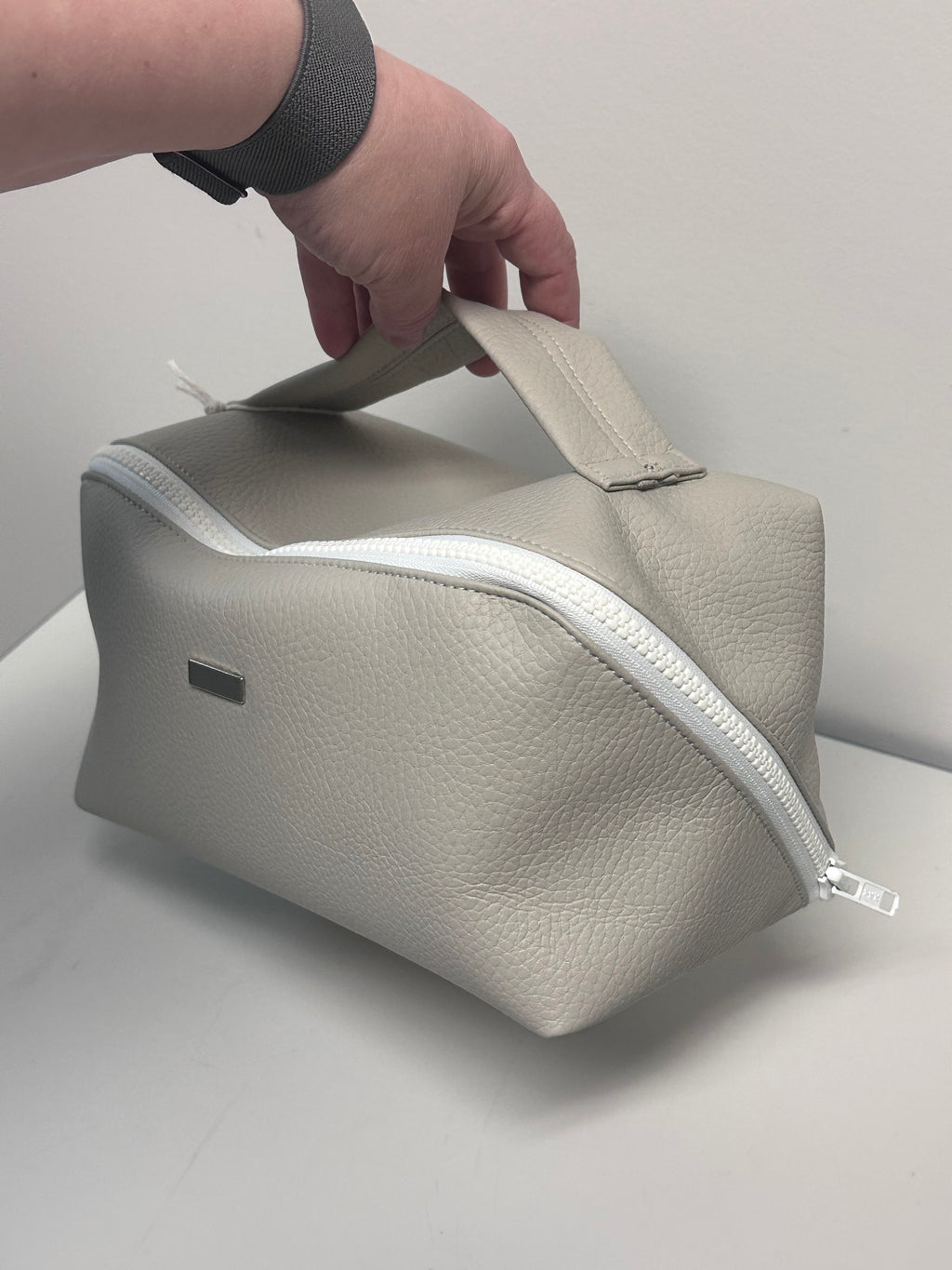Hand holding a grey faux leather pouch with a zipper on a light gray background