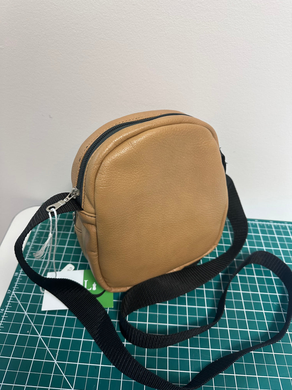 Tan crossbody bag with a black strap on a white surface with a grid pattern.