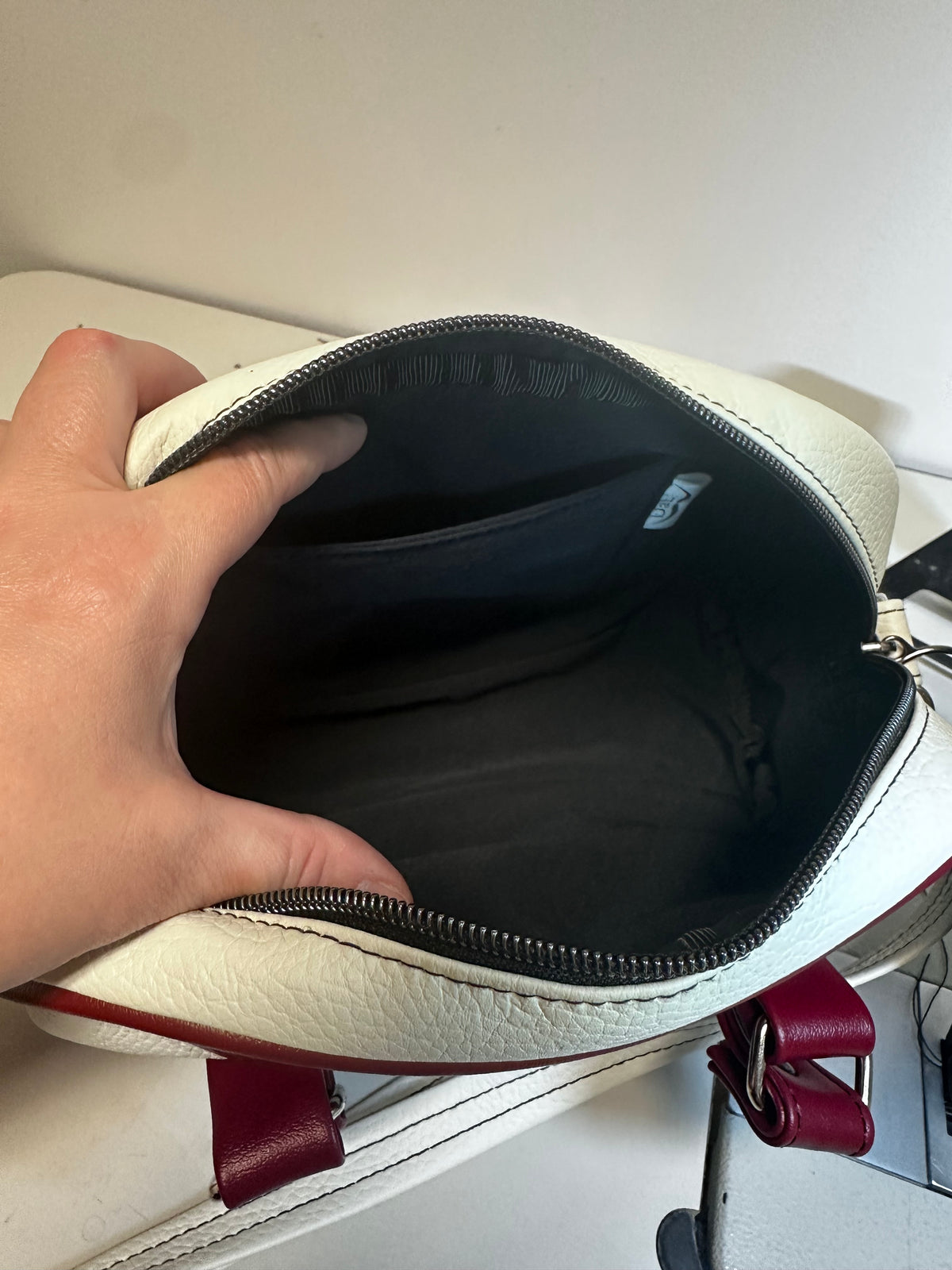 A close-up of a hand holding open a white textured leather pouch or small bag, showing a dark gray interior lining and zipper. A red leather strap is visible at the bottom right against a light neutral background.