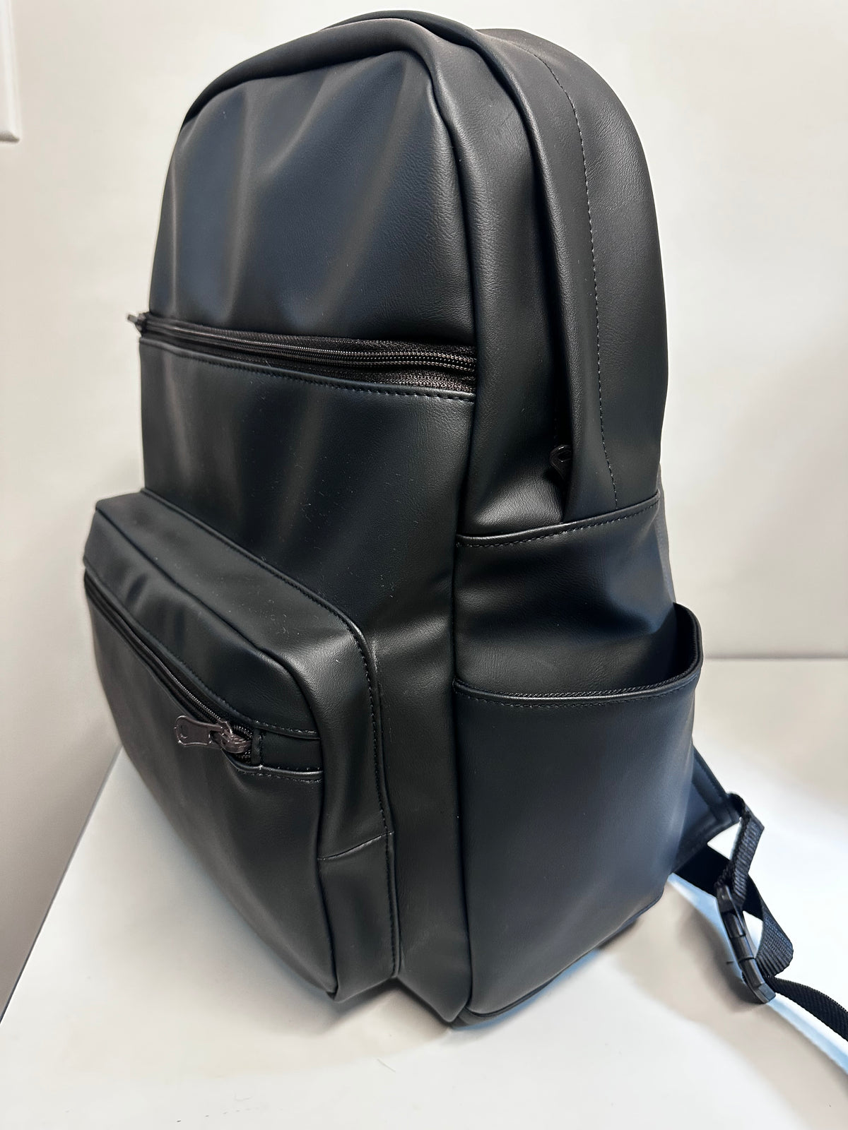 A black faux-leather backpack with multiple zippered compartments and side pockets, shown from a three-quarter angle on a white surface against a white background.
